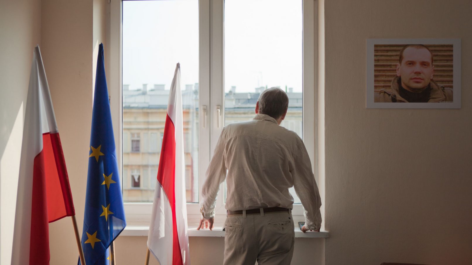 Mann steht am Fenster und sieht nach draußen.Neben ihm drei Fahnen, die der Europäischen Union und weiß-rot von Belarus.