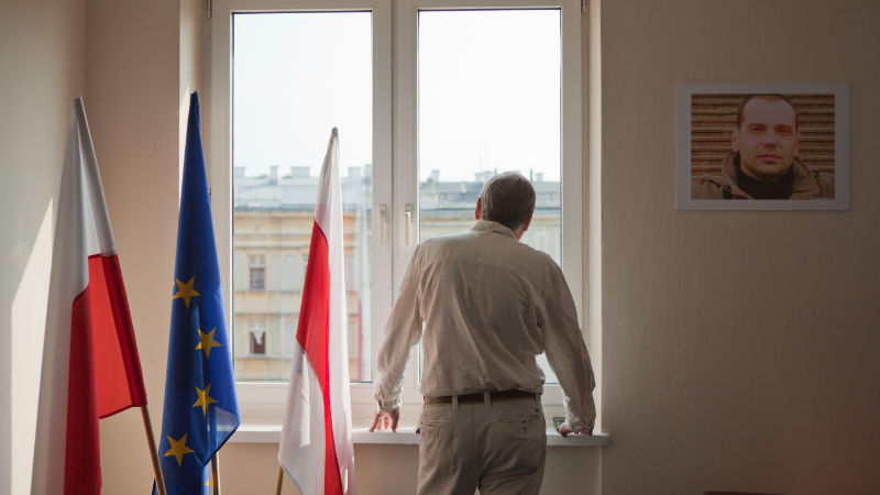 Filmstill. Mann in Hemd und heller Hose steht mit dem Rücken zur Kamera am Fenster. Links neben ihn drei Flaggen, zwei weiß-rot, eine europäische in der Mitte. Recht neben ihm hängt ein Bild eines Mannes an der Wand.