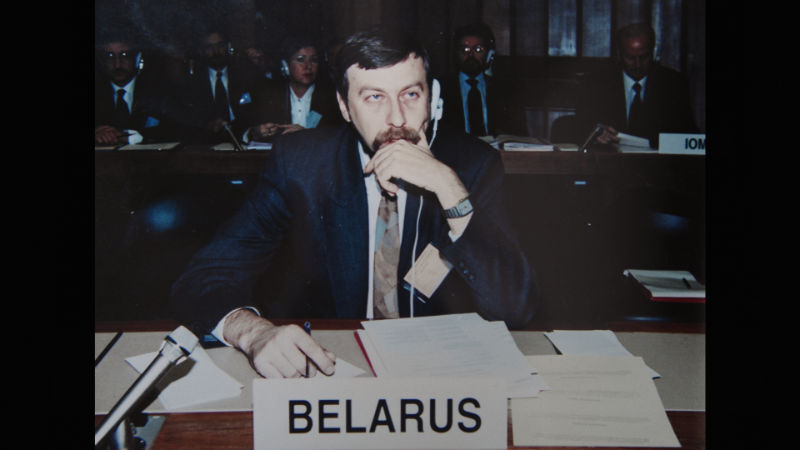Ein Mann im Anzug mit Kopfhörern sitzt hinter einem Konferenztisch. Vor ihm ein Aussteller mit der Aufschrift: Belarus