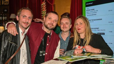 Foto: Drei Männer und eine Frau stehen an der Theke des Filmfest Bremens, gekennzeichnet von grünen Programmhefen.