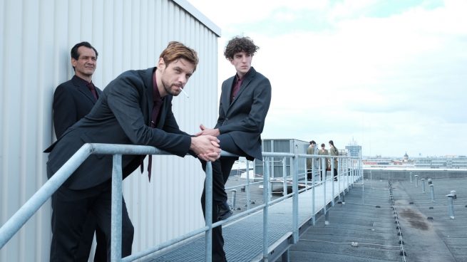 Drei Männer im Anzug stehen auf einem Dach vor einer Containerbaracke und starren nach links. Im Hintergrund drei Frauen im Kreis.