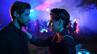 Eine Frau mit hochgesteckten Haaren legt einem Mann im Anzug eine Hand auf die Schulter. Im Hintergrund sind die Lichter und Nebel einer Partyszene.