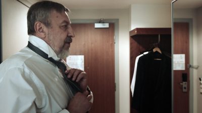 Ein Mann Ende 60 bindet sich im Gang eines Hotelzimmers seine Krawatte.