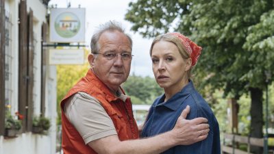 Ein Mann (Uwe Preuss) und eine Frau (Nina Proll) stehen unter Bäumen vorm Vereinsheim eines Kleingartenvereins.